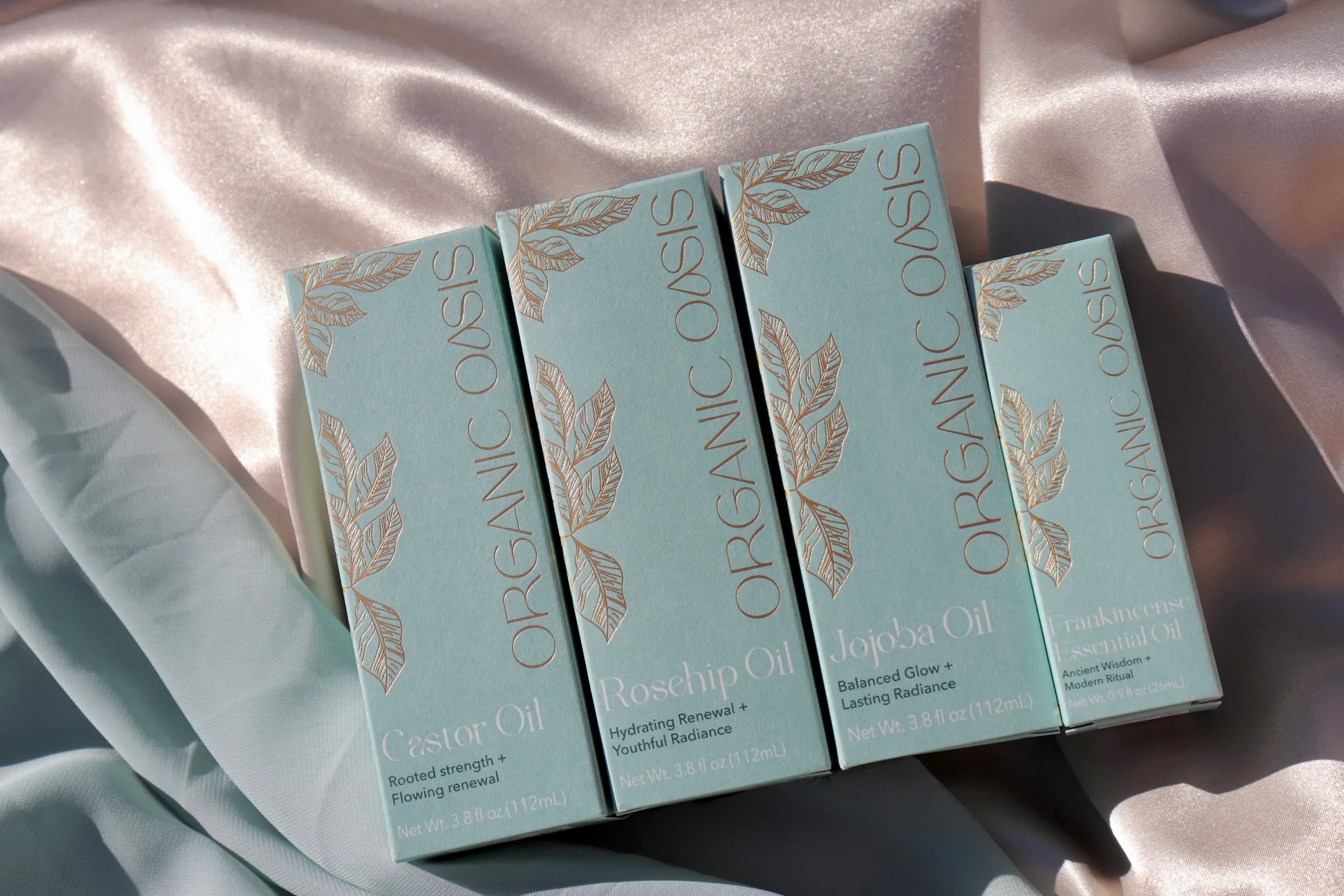 Luminous Collection, boxes of Organic Oasis body oils arranged diagonally on draped satin fabric. The boxes are a pale mint green with gold foil botanical leaf designs and gold lettering, showing the varieties: Castor Oil, Rosehip Oil, and Jojoba Oil.